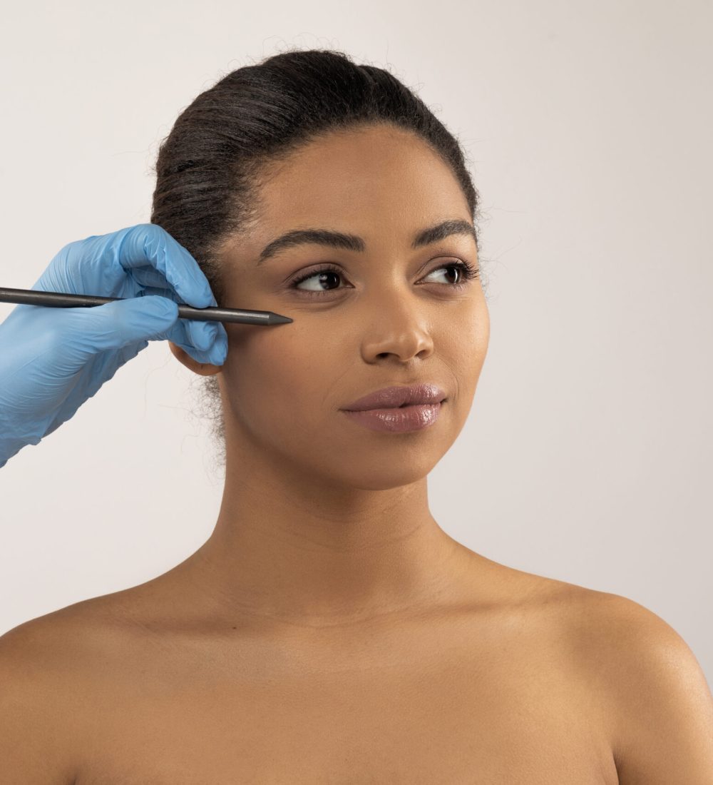Aesthetic medicine, injection cosmetology concept. Plastic surgeon hands in medical gloves holding pen, making preparation lines on beautiful young black woman face, isolated on grey background